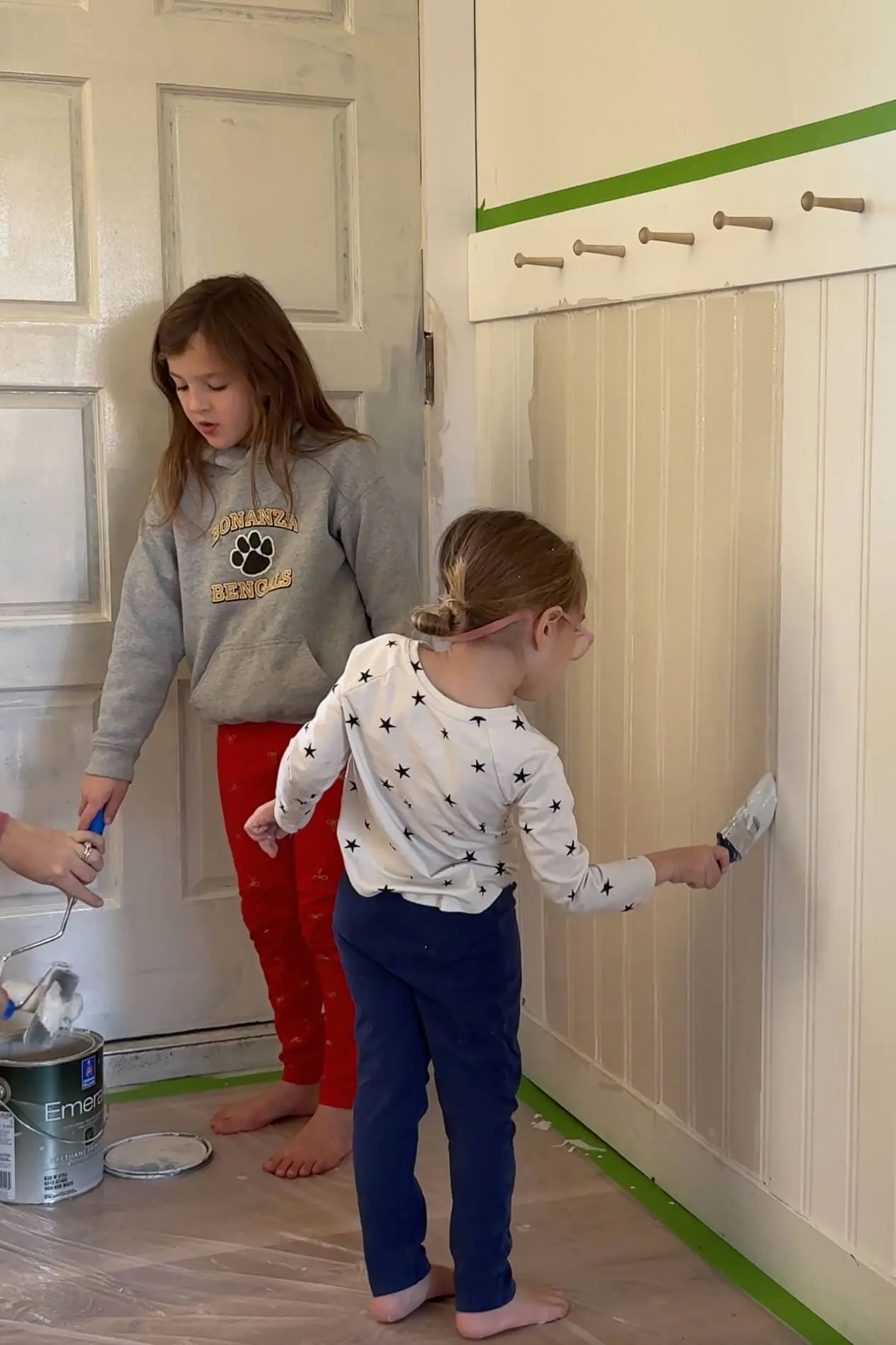Two young children helping with a DIY painting project, one carefully using a paintbrush to coat beadboard paneling while the other observes. A can of paint and a roller rest on a plastic-covered floor in a freshly updated room with painter’s tape lining the wall.